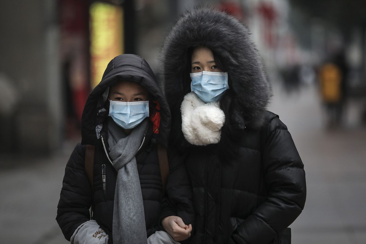 Two women in winter coats with hoods stand outside on a street in China wearing breathing masks.