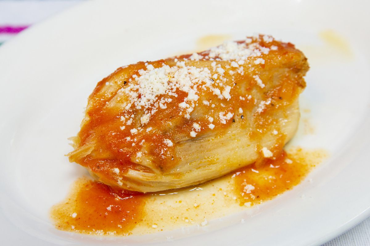 A white plate with a tamale wrapped in a corn leaf and topped tomato sauce and shredded cheese