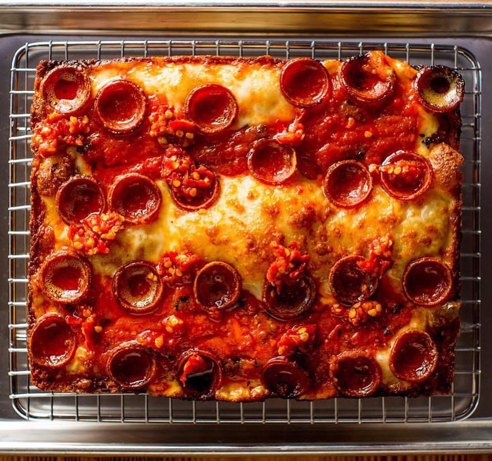 A top-down view of a rectangular Detroit-style pizza covered with cheese, stripes of red sauce, and pepperonis.