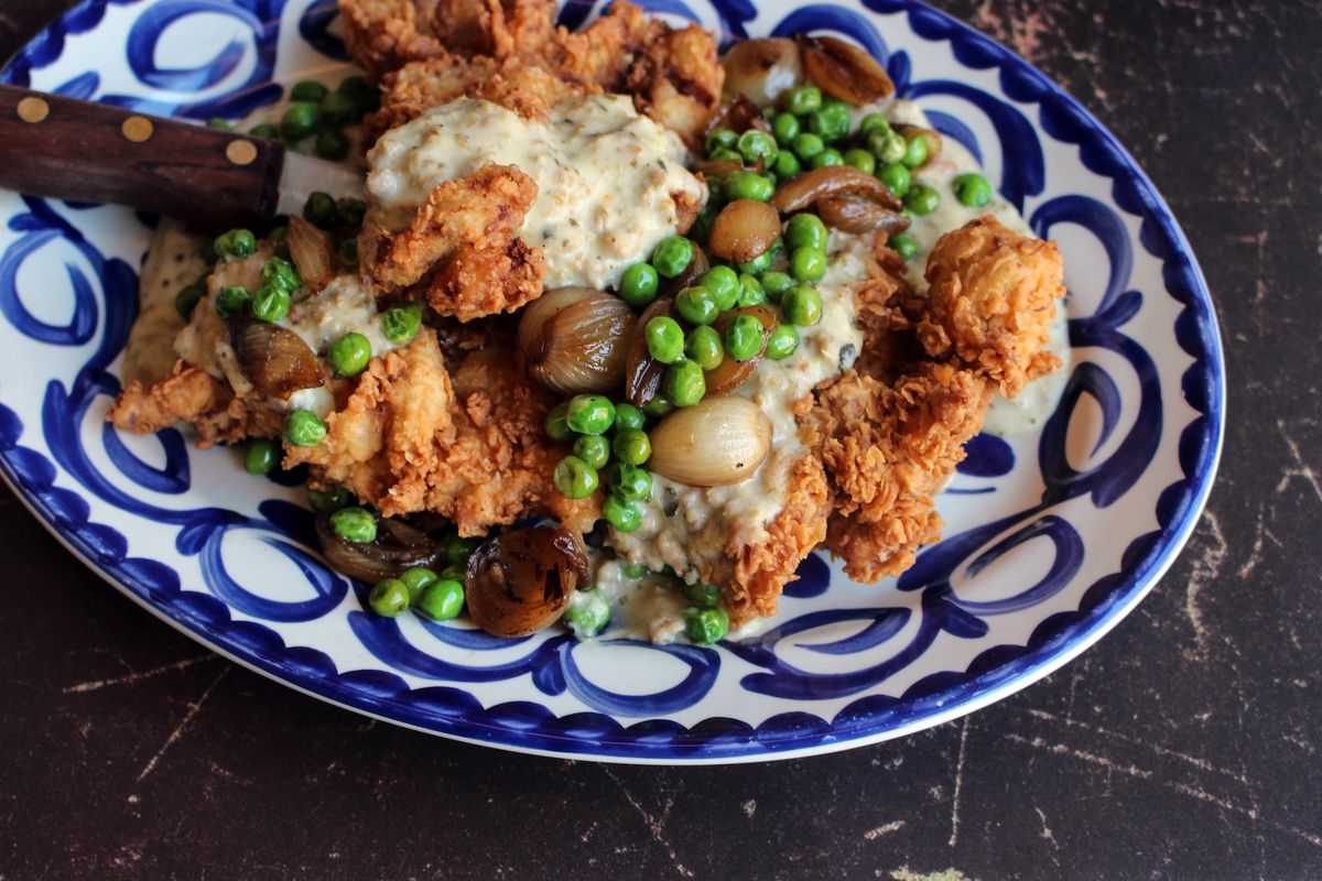 A plate of fried chicken covered with white gravy, peas, and pearl onions