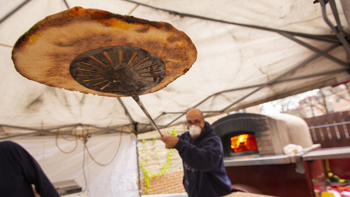 A man lifts a pizza out of an wood-burning oven outside.