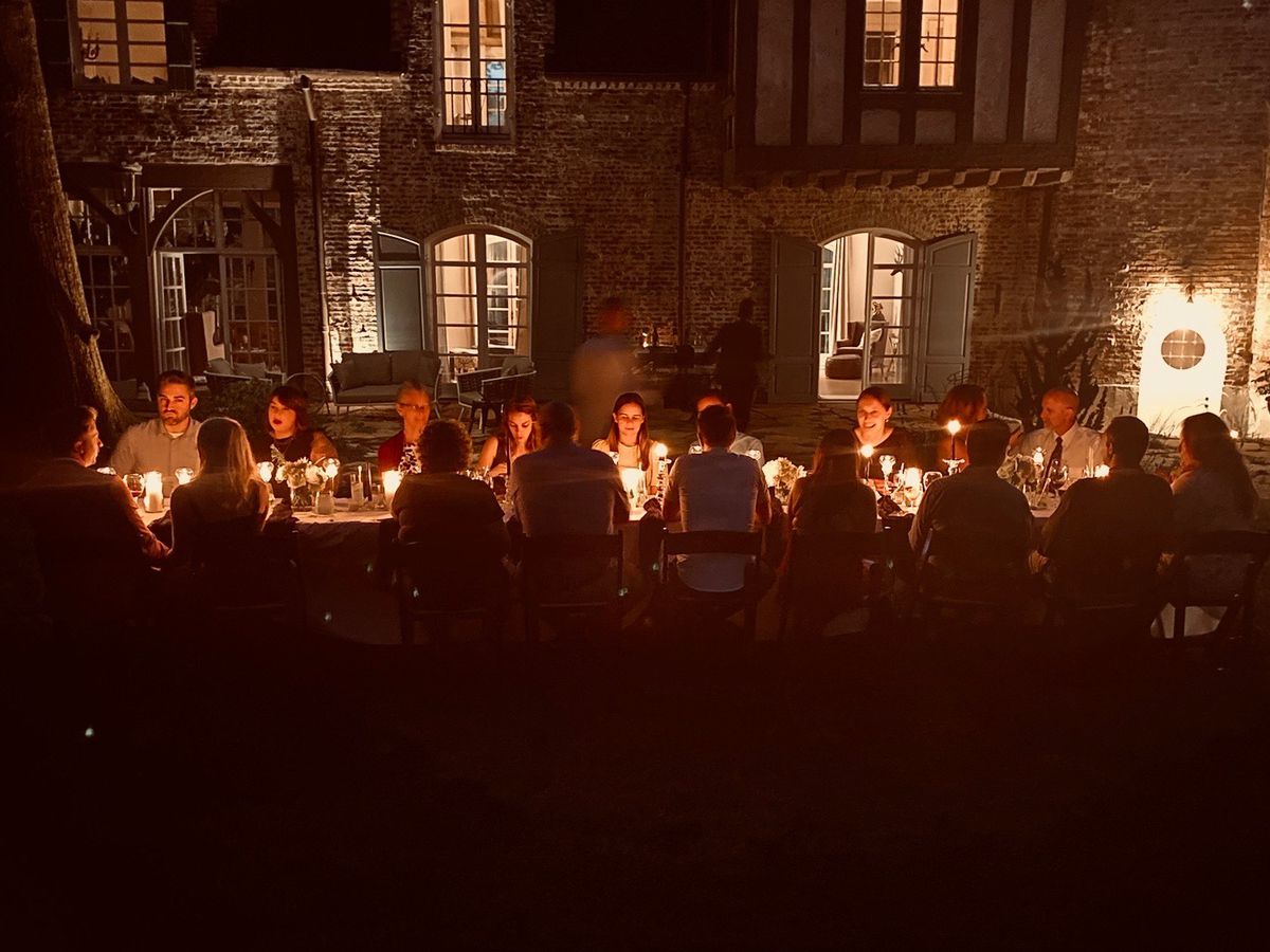 Diners seated at a long candlelit table in front of a large mansion. 