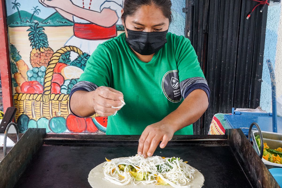 An Indigenous woman cooks up a cheese and squash blossom-filled tortillas on a steel grill on the sidewalk.