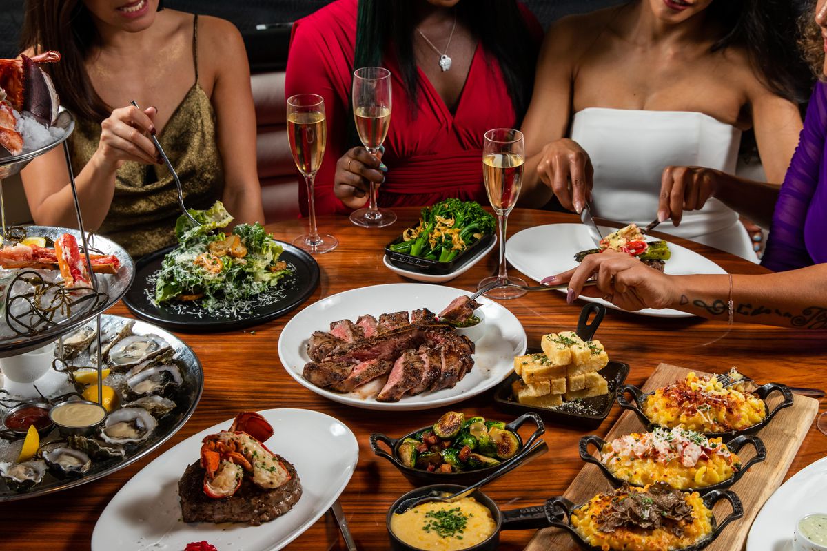 Four women sit at a table loaded down with steaks and steakhouse foods.
