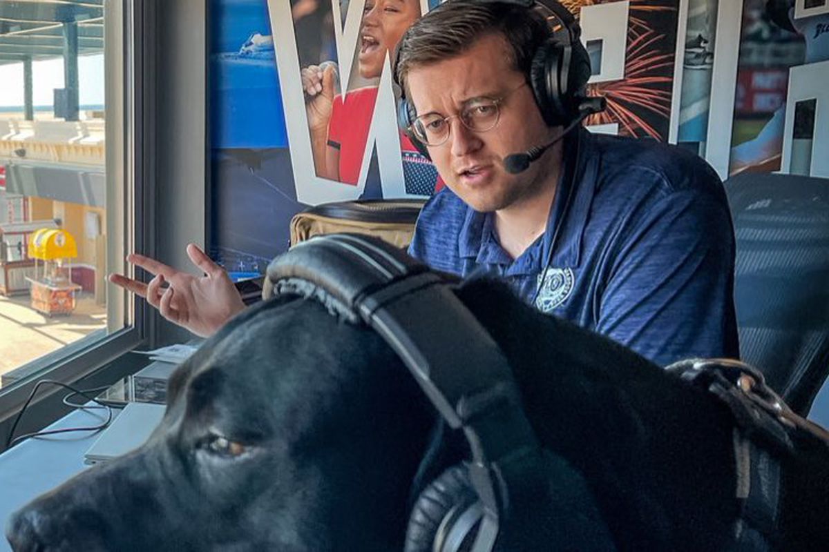 Erik Bremer sits in the broadcast booth at Blue Wahoos Stadium alongside a large dog wearing headphones