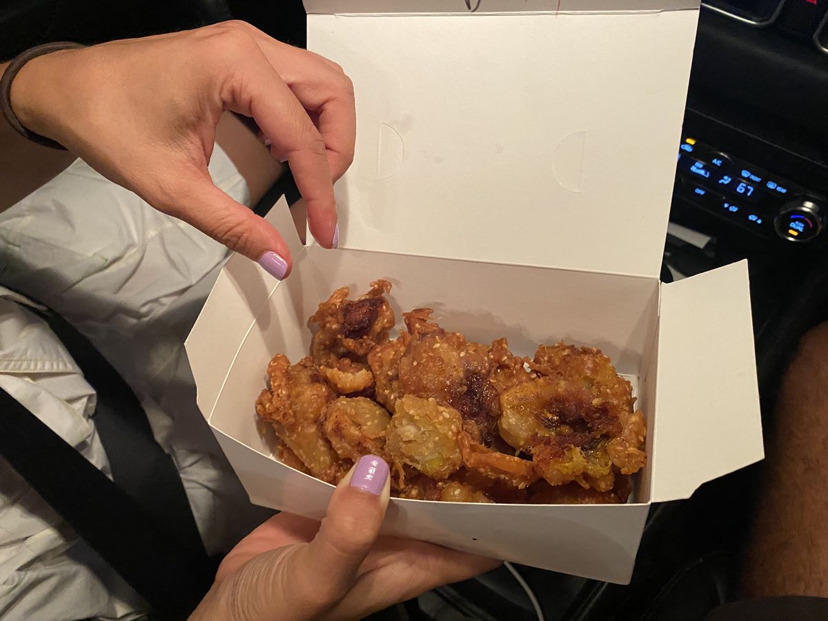 A hand reaches into a a white takeout box containing golden-fried chunks of banana.