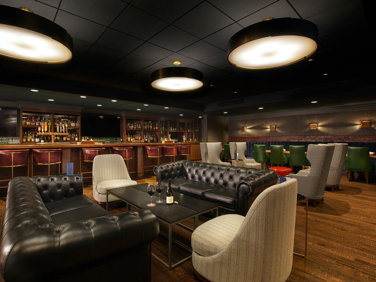 A darkened bar interior with plush leather couches and chairs, and a dimly lit bar with lots of bottles