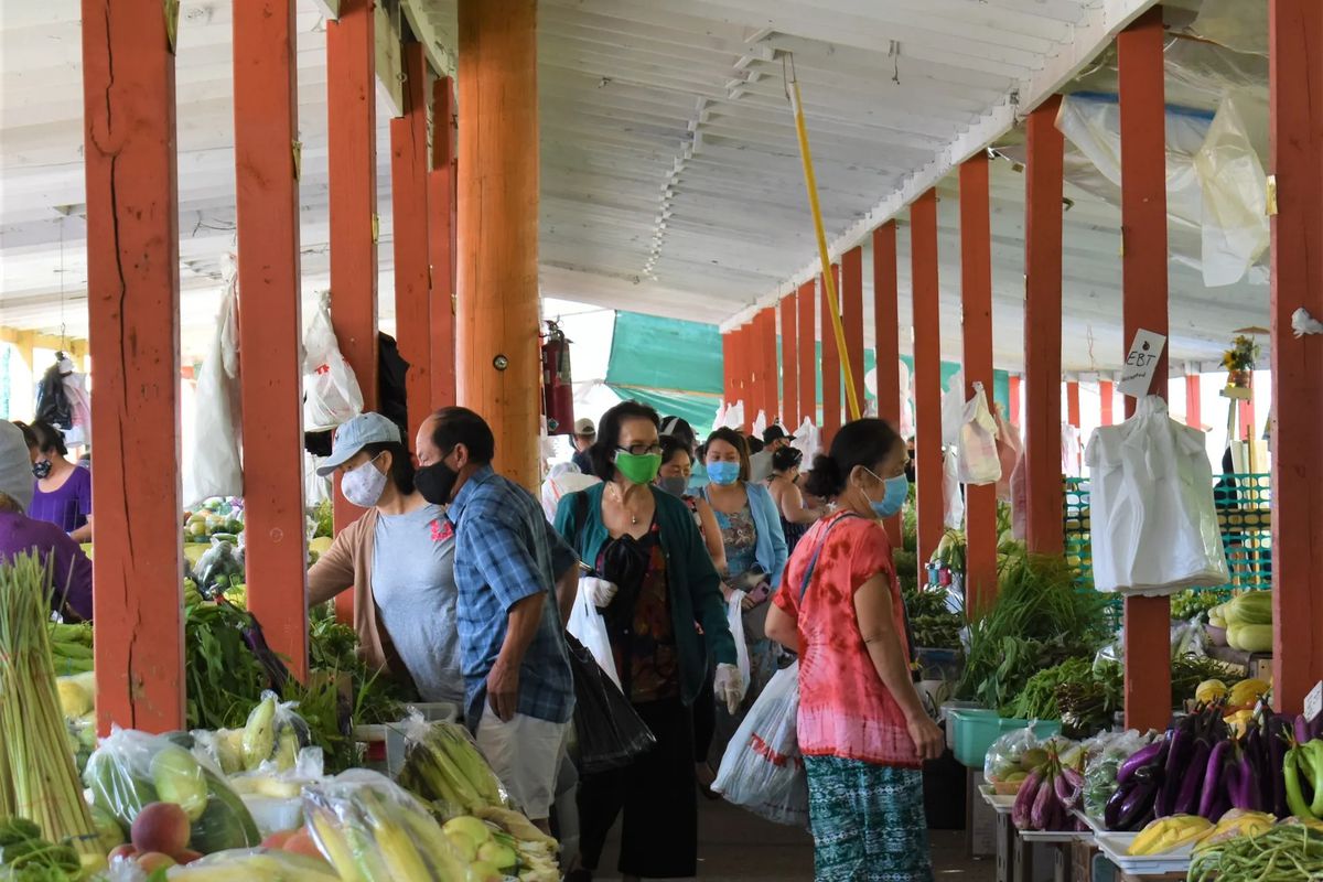 People wearing hygenic masks shopping for fruit and vegetables in a covered space with wooden pillars and a white ceiling. 