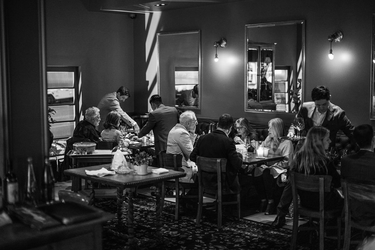 Diners and staff of a fine dining restaurant in a dimly lit dining room, in black and white.