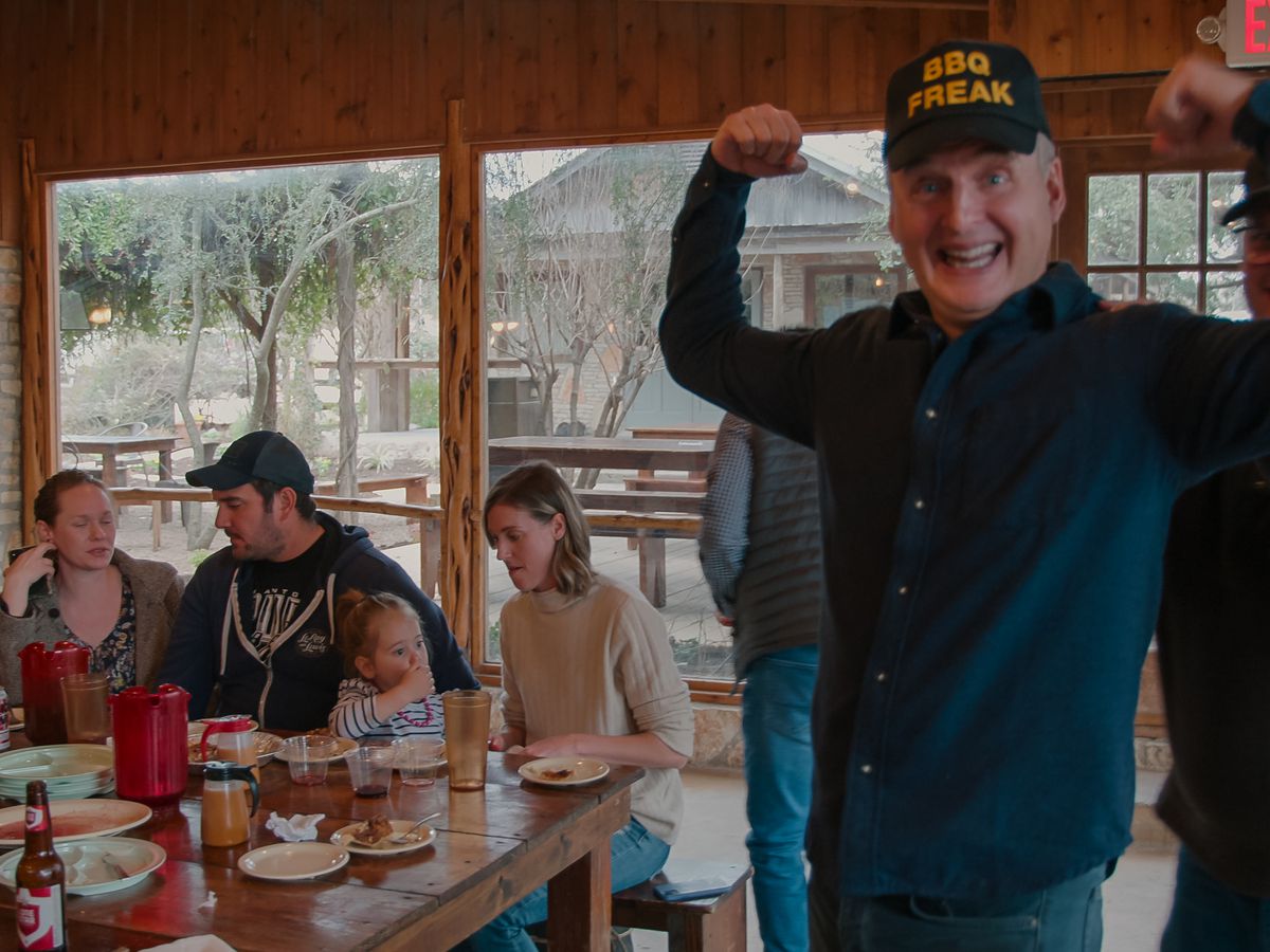 A man with his arms up wearing a baseball cap that reads “BBQ Freak” with people dining behind him.