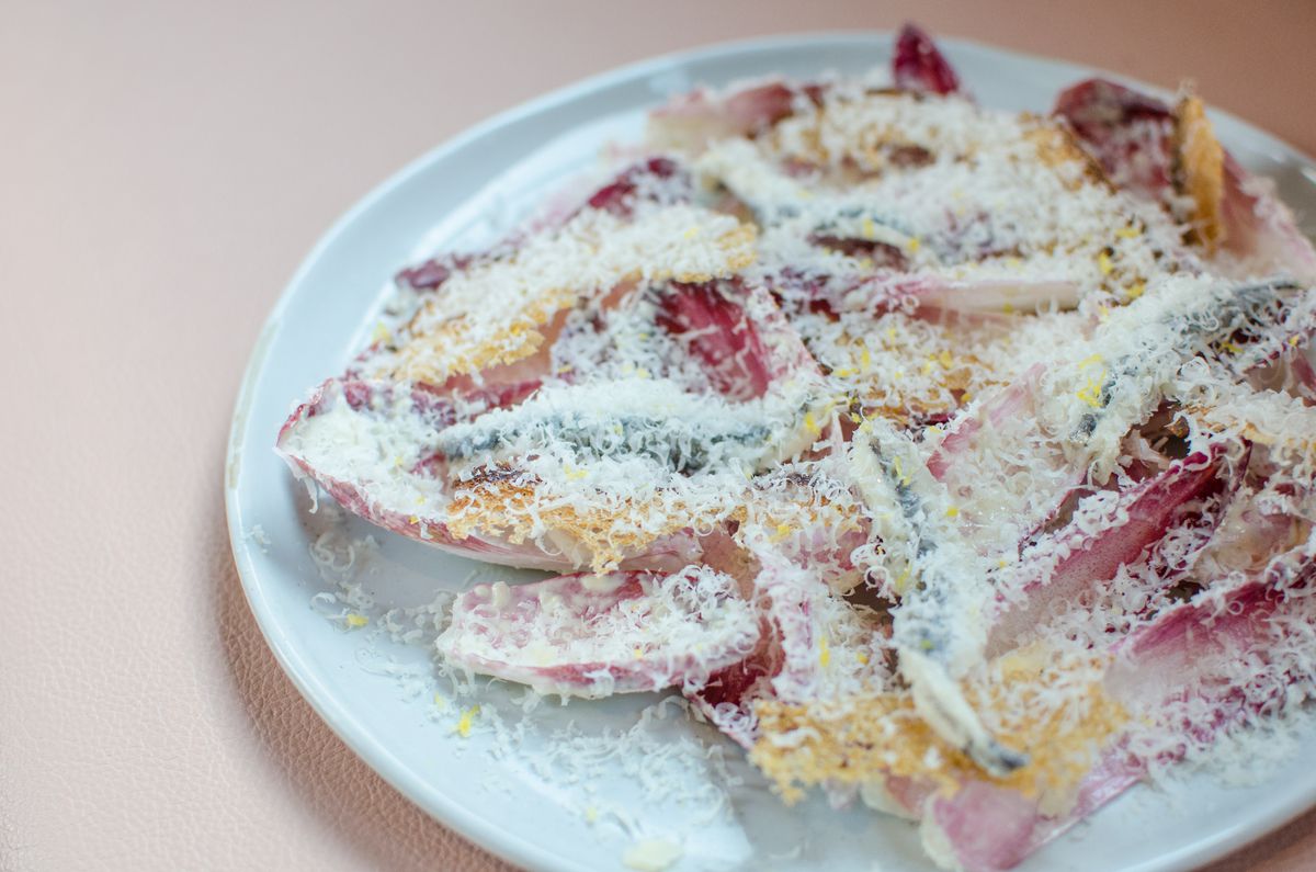 A plate full of red endives topped with fat silver hunks of anchovy and tons of shaved cheese sits on a pale pink background.