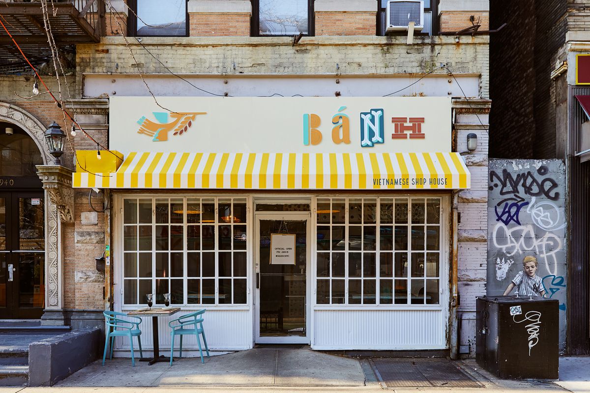A colorful restaurant exterior with a yellow and white striped awning and white framed windows, with a teal blue set of chairs and a table set up outside