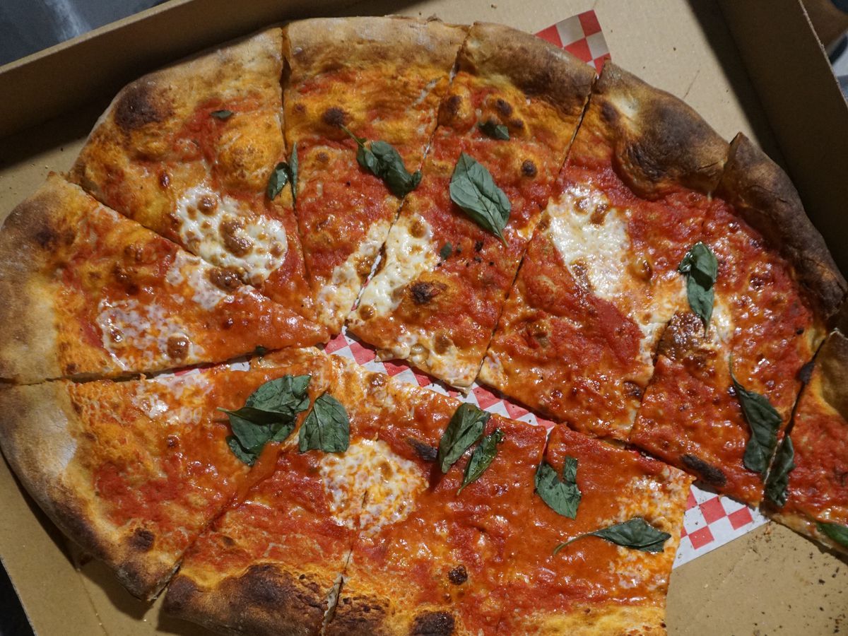 An oval-shaped pizza cut into wedges, topped with tomato sauce, globs of cheese, and leaves of basil. The pizza is in a cardboard box with one slice missing