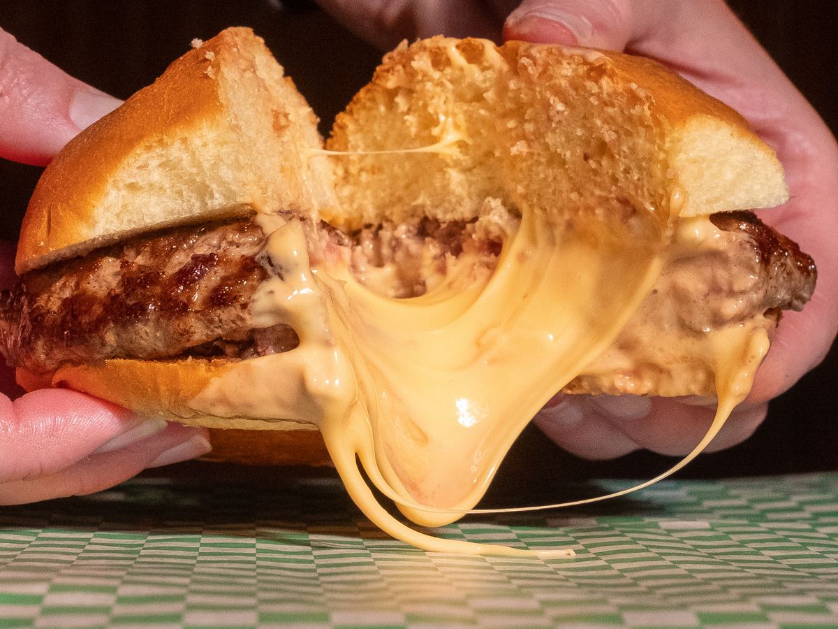 A juicy lucy cut in half with prodigious amount of cheese spilling from the center as two hands pull it apart. 