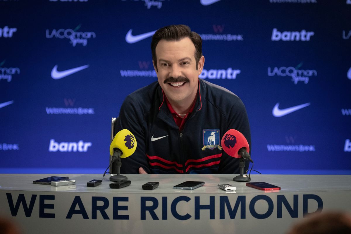 Ted Lasso (Jason Sudeikis) sitting and smiling in front of press mics at a press conference