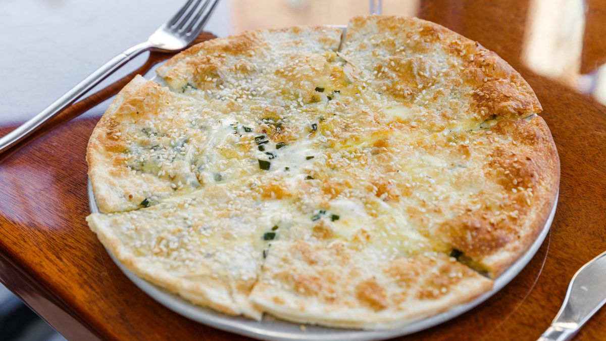 A round, sesame-studded flatbread is cut up into slices on a big plate on a reddish wooden table.