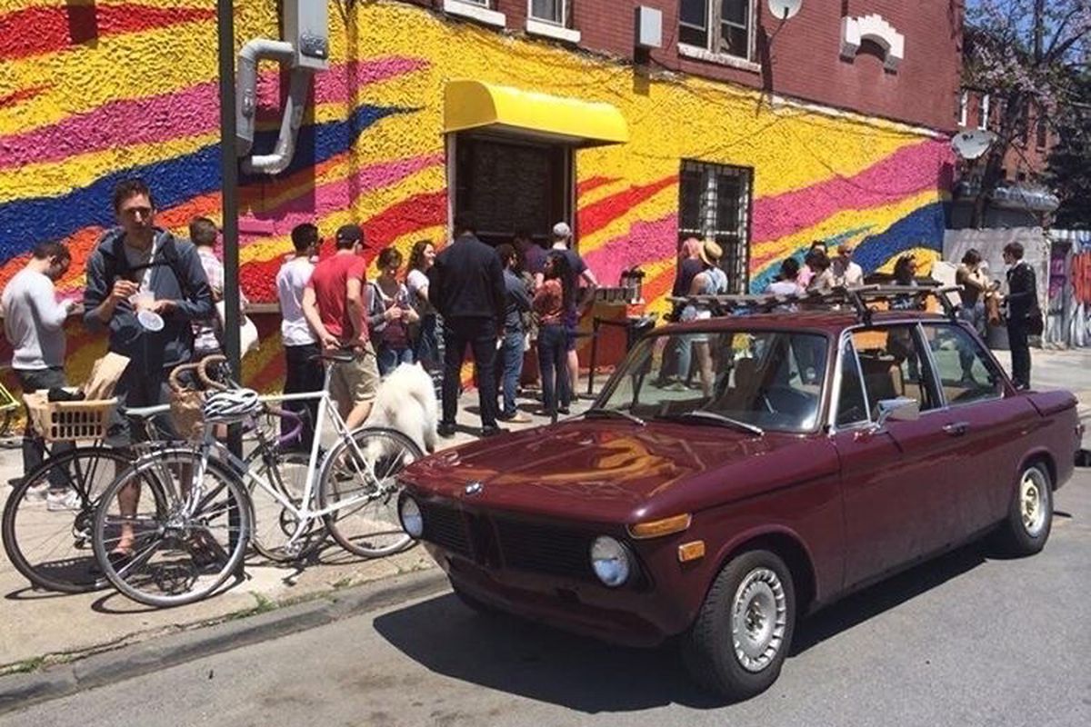 A crowd of people stands in front of a brightly painted wall with bikes and a red car visible in the street