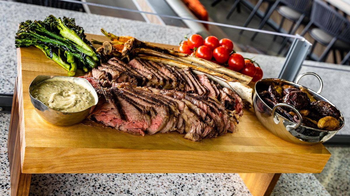 A steak is served on an elevated butcher block with a silver tray of roast potatoes, a silver bowl of sauce, tomatoes, and roast broccoli.
