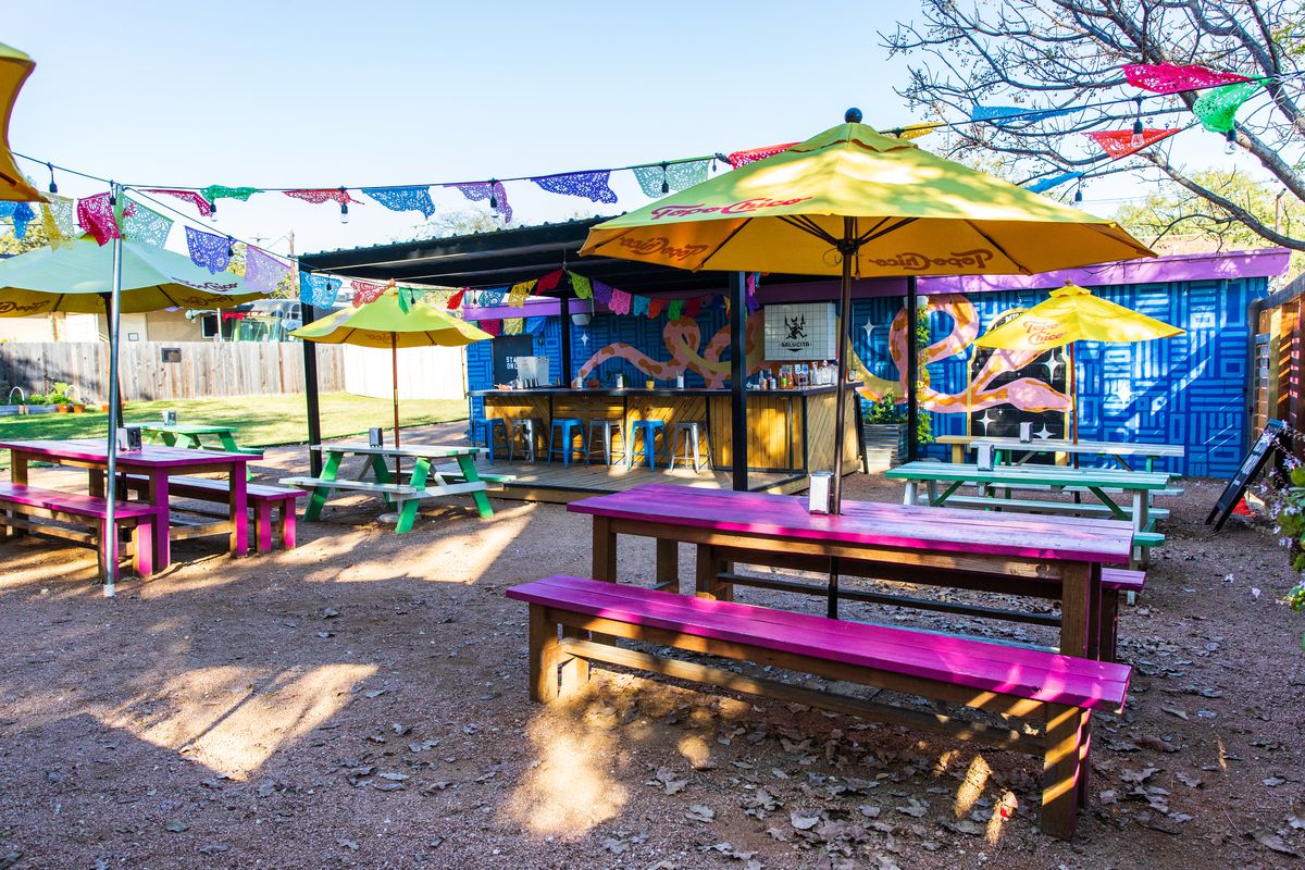 A restaurant outdoor area with purple and green picnic tables, yellow umbrellas, in front of a colorful blue building.