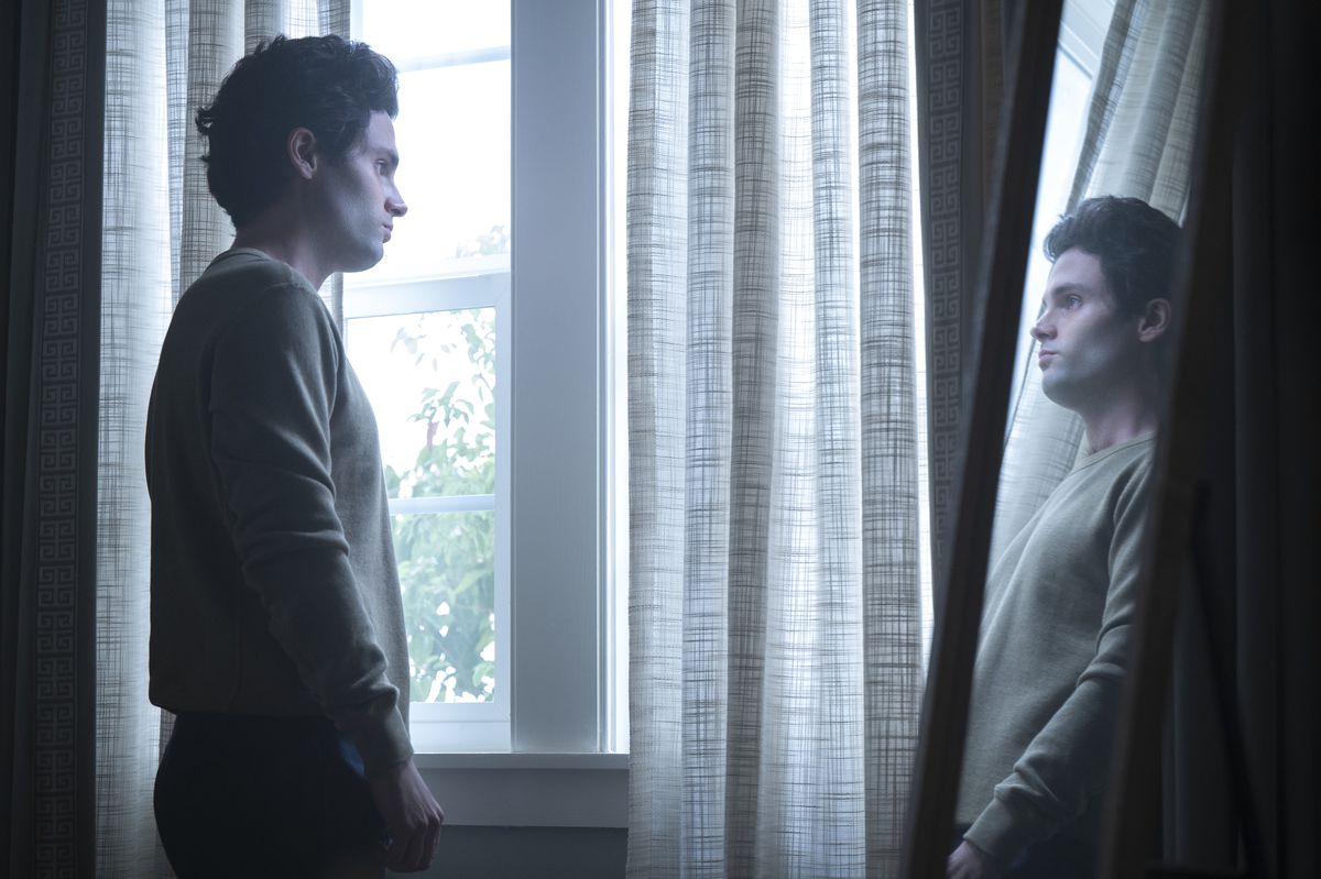 PENN BADGLEY as JOE GOLDBERG stands looking out a window but reflected in a mirror in You