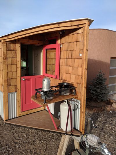An exterior area on a travel trailer. There is a red door, an outside table with cooking tools, and wood shingles on the walls. 