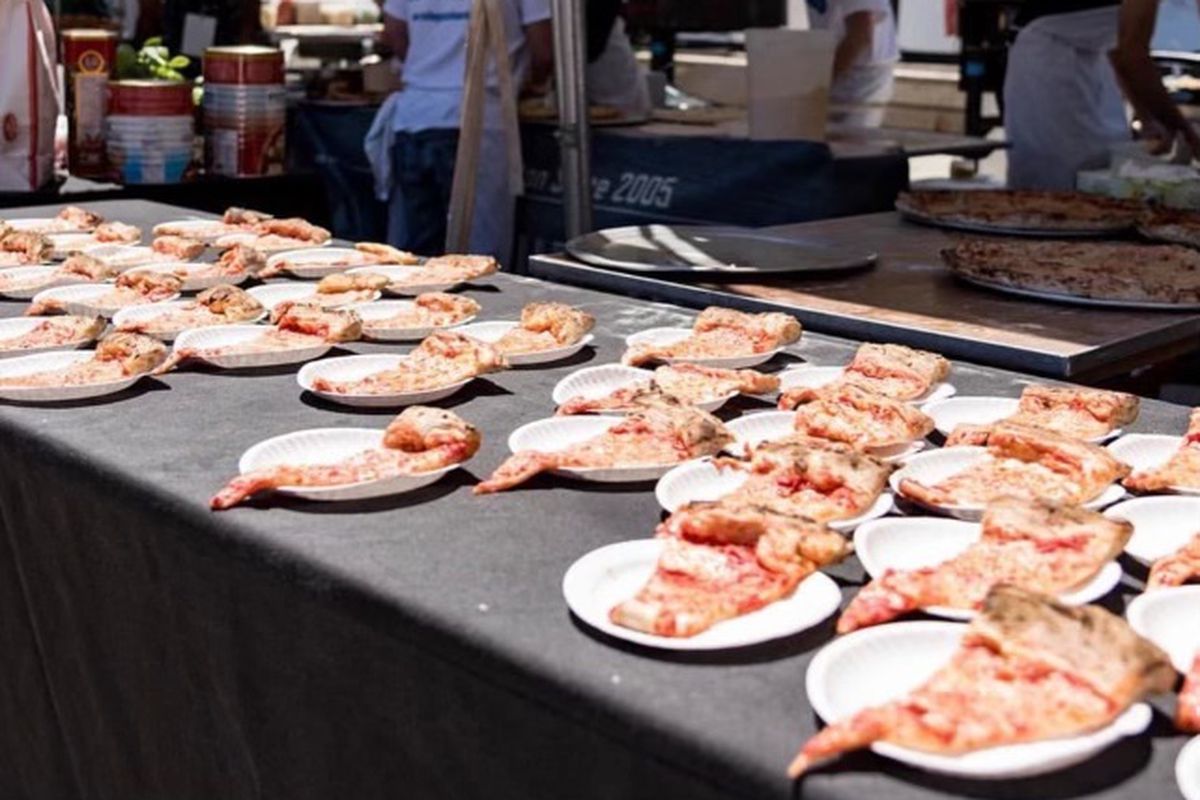 Slices of pizza lined up on individual white paper plates at a booth at a past Boston Pizza Festival event.