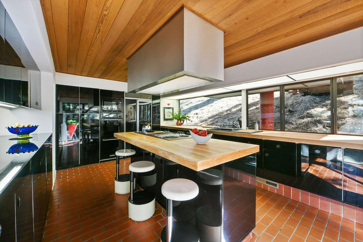 A kitchen with a wood paneled ceiling, a kitchen island with three white circular stools, and glossy black cabinets and drawers.