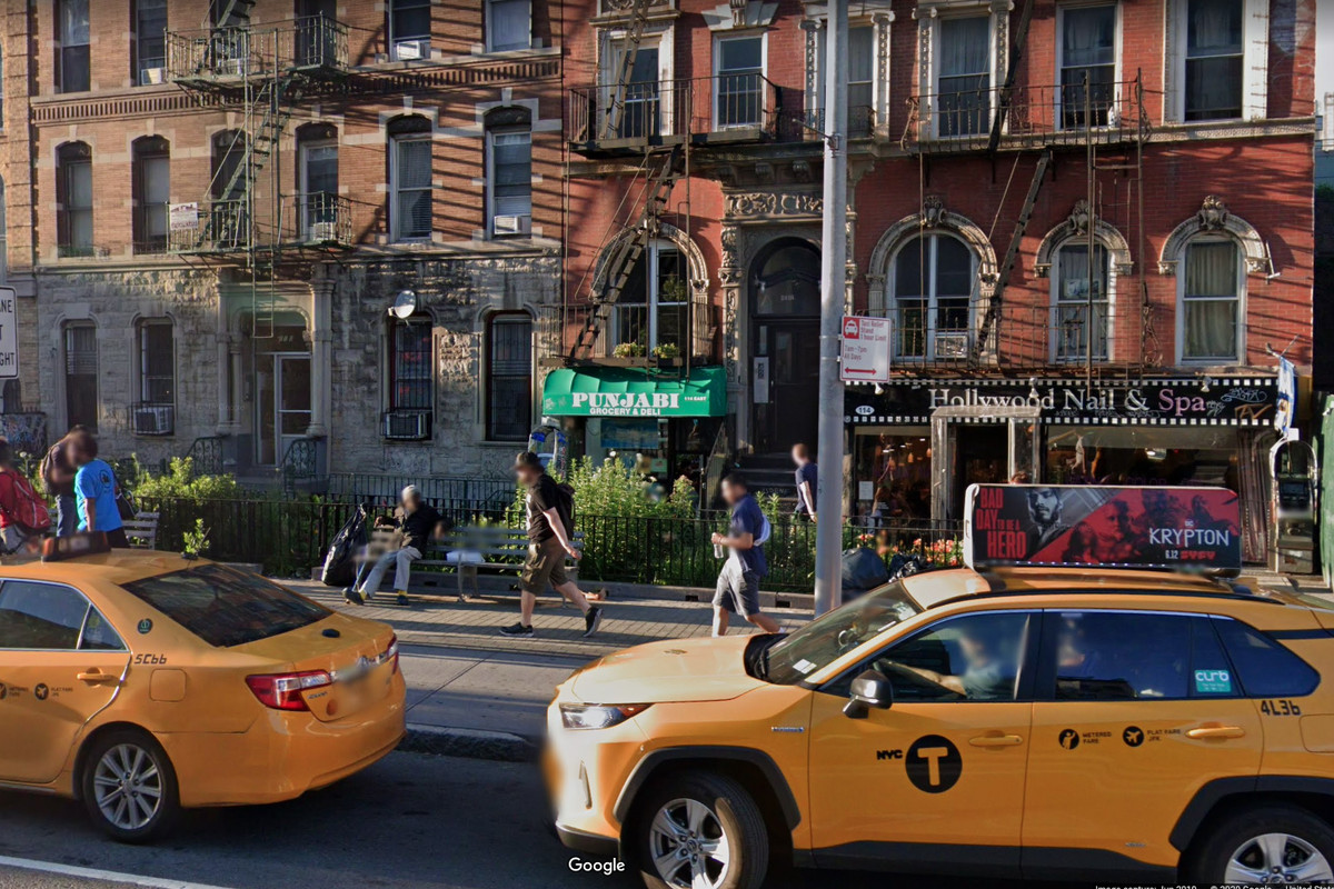 An exterior photo of the deli with a green awning out front and two yellow taxi cabs in the foreground