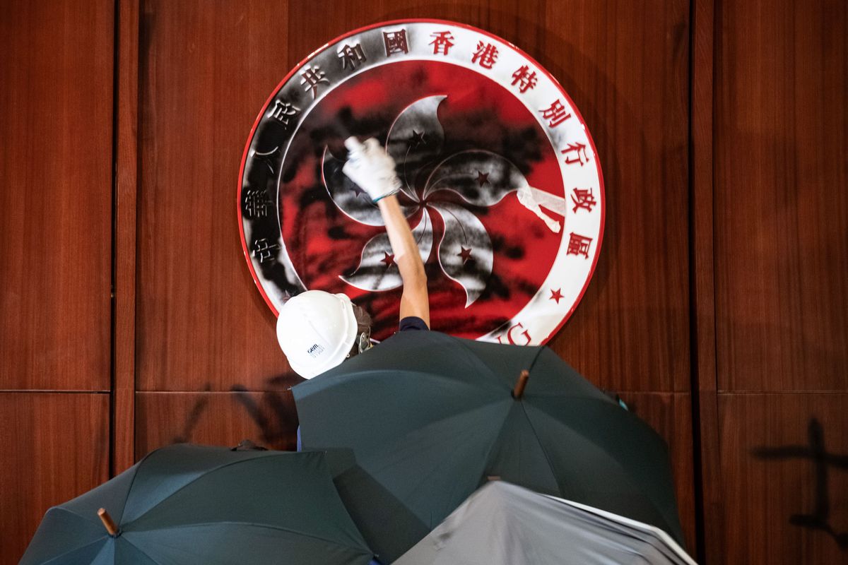 A protester defaces the Hong Kong emblem after protesters broke into the legislative chamber on July 1, 2019. The protester is framed by umbrellas, a symbol of Hong Kong’s past protest movement.