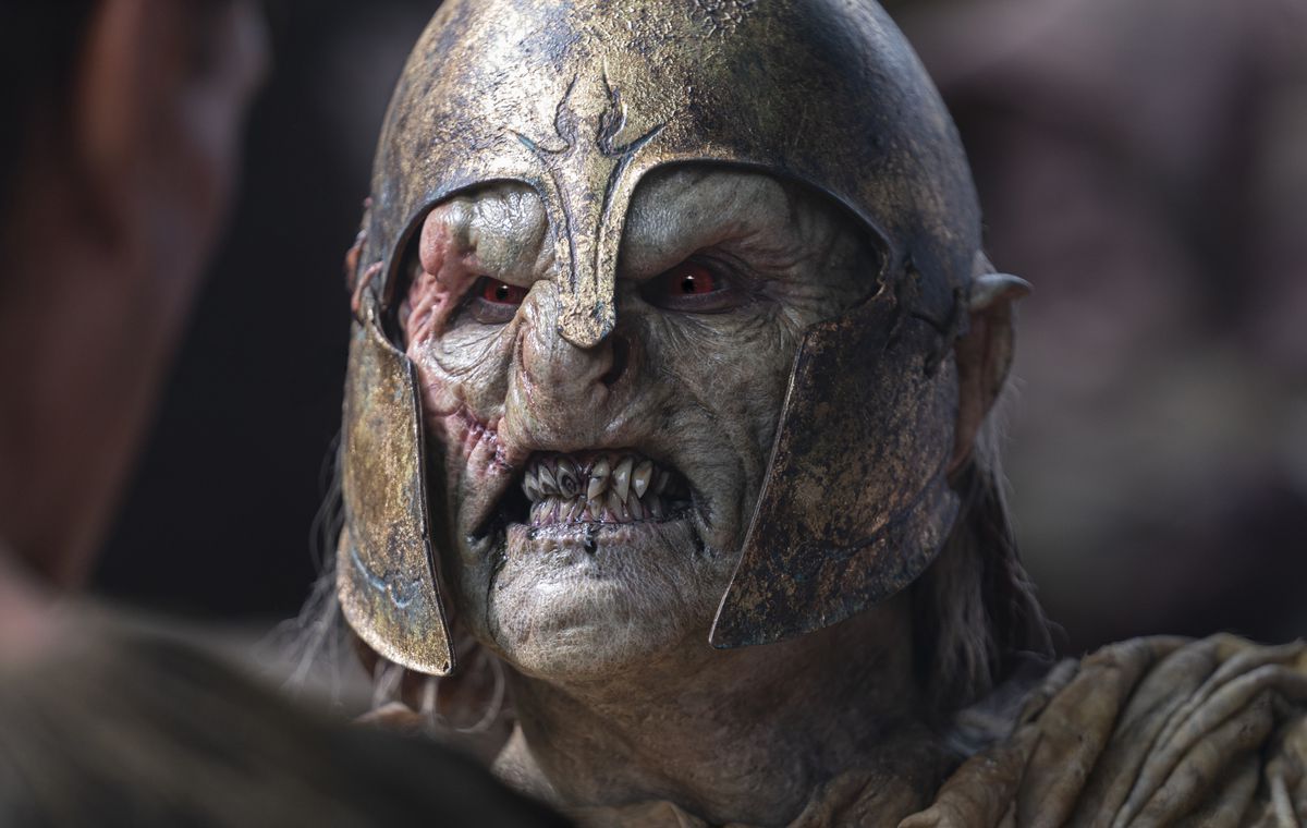 A close-up of an orc in The Lord of the Rings: The Rings of Power. The creature has sallow skin and is wearing a rusted elven helmet as it snarls.