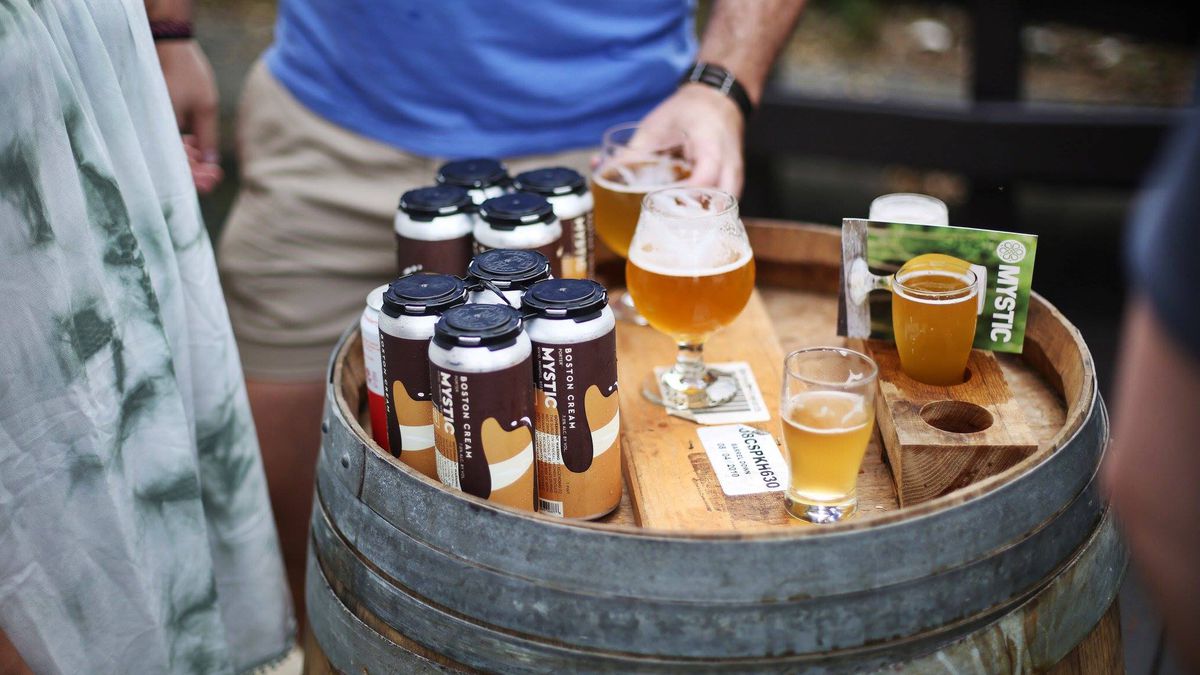 A glass of beer and beer cans stand on top of a barrel; people are standing around the barrel, although only some legs and arms are visible.