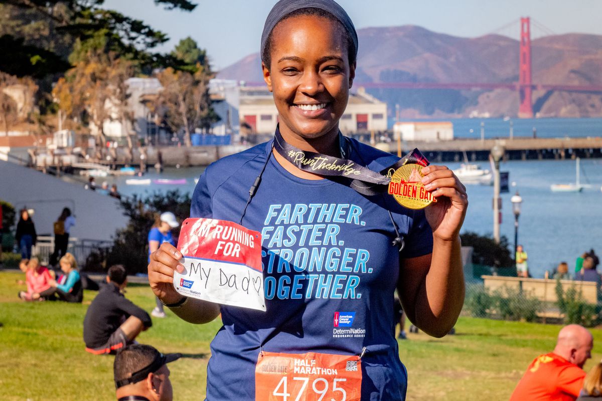 A Black woman shows off a gold medal and a race bib that reads “Running for My Dada,” having completed a half marathon near the San Francisco Golden Gate Bridge. 