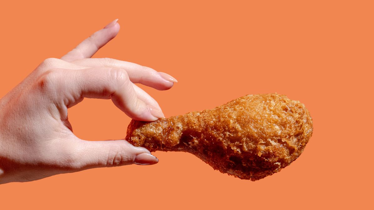A hand with painted nails holds up a piece of fried chicken against an orange background.