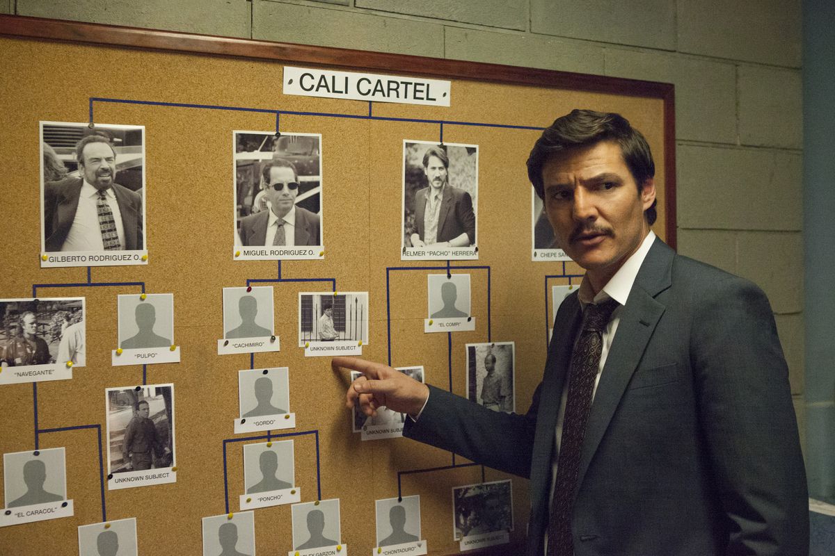 An actor playing a detective on the Netflix show “Narcos” stands in front of a corkboard covered in a relationship tree of photos labeled “Cali Cartel.” 