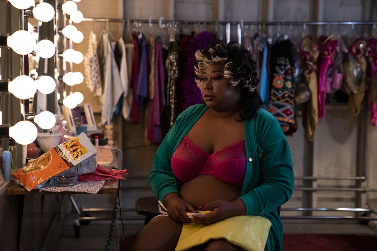 Tammé sits in a dressing room looking unfocused — she is wearing a pink bra and open teal robe. Her hair has silver streaks and is curled.