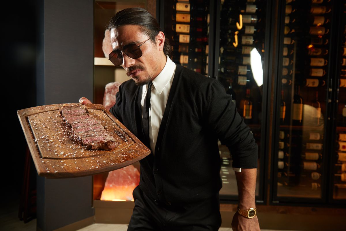 Salt Bae lovingly holding a chopping board topped with a steak, while wearing a suit, tie, and sunglasses