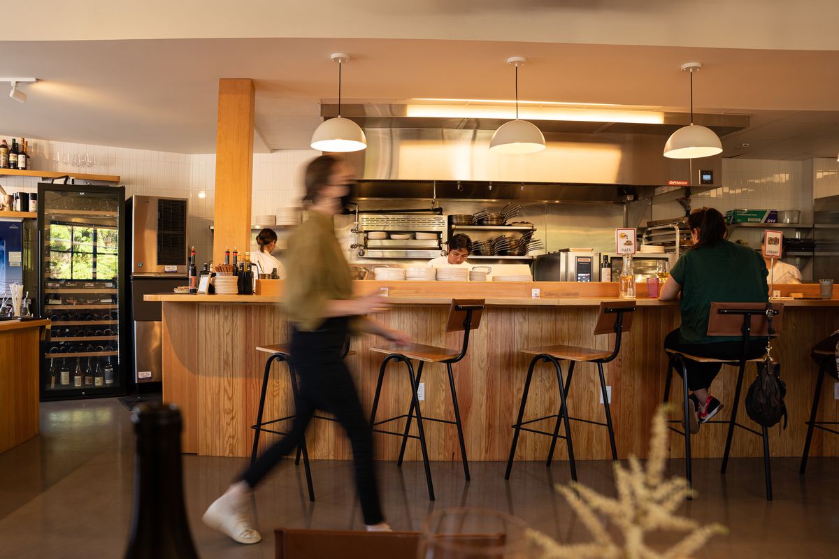 A restaurant dining room with a wood counter in front of a kitchen and a person is moving blurred through the space.