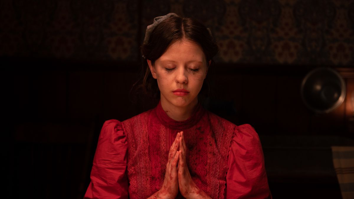Mia Goth as Pearl in Ti West’s Pearl, wearing a red dress and with her hands folded in prayer