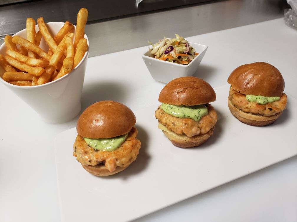 Three salmon sliders sit atop a white plate, along with slaw and French fries.