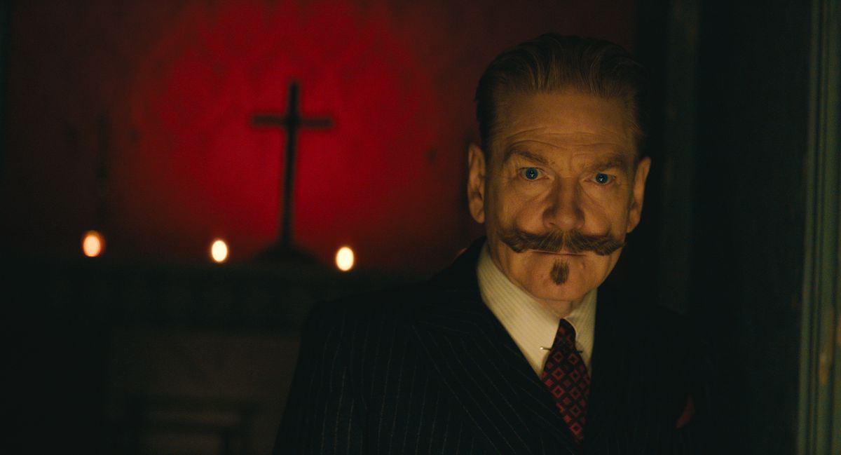 Kenneth Branagh’s Hercule Poirot, standing in a dark room. A cross hangs on the wall behind him.
