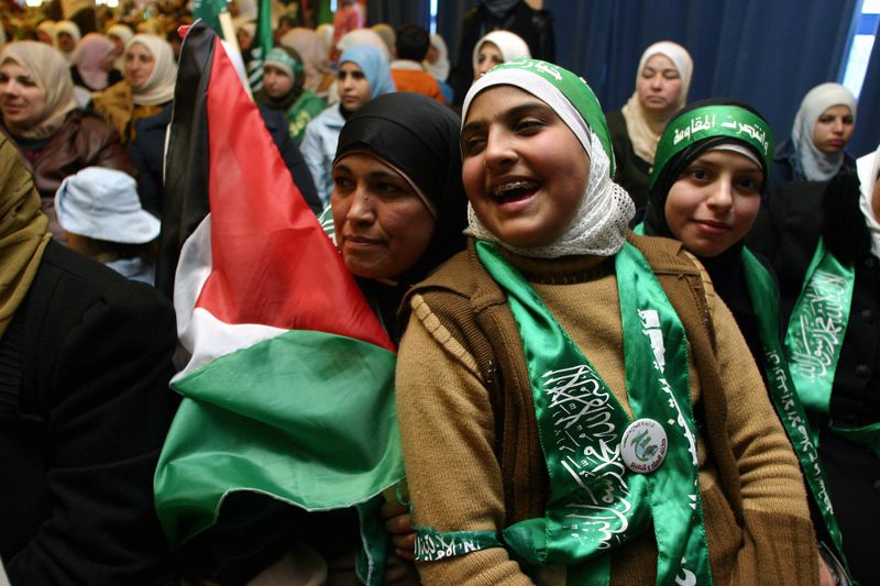 A young woman in a white hijab with a green Hamas headband and a green Hamas scarf cheers as she leans against an older woman who holds a Palestinian flag. Other women, many with Hamas gear smile behind them. 