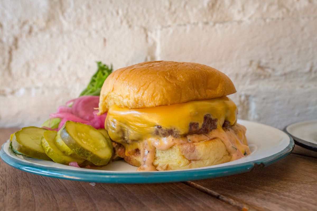 A photo of a burger placed on a blue plate on a wooden table against an exposed brick wall
