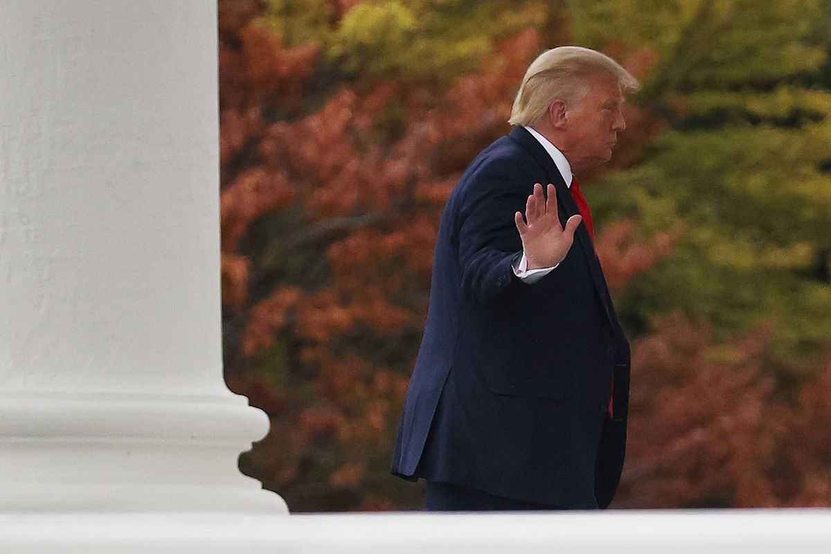 President Donald Trump walking near the White House looks away but holds up one hand with his palm toward the camera.