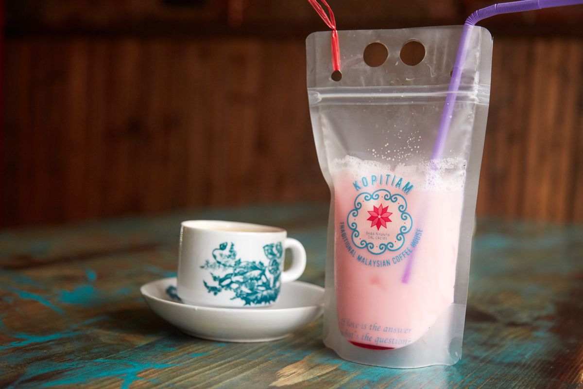 A clear plastic bag, labeled with the red and light blue logo of NYC’s Kopitiam, is full of light pink liquid. It sits on a stripped wooden table next to a white teacup and saucer decorated with blue flowers. 
