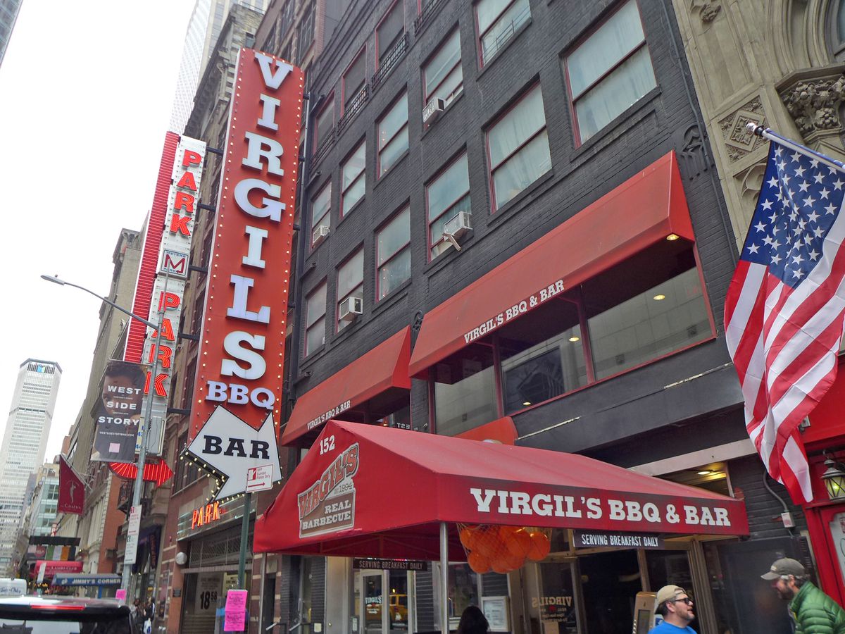 A barbecue with red signage and skyscrapers all around it and an American flag hanging down on the right of the picture.