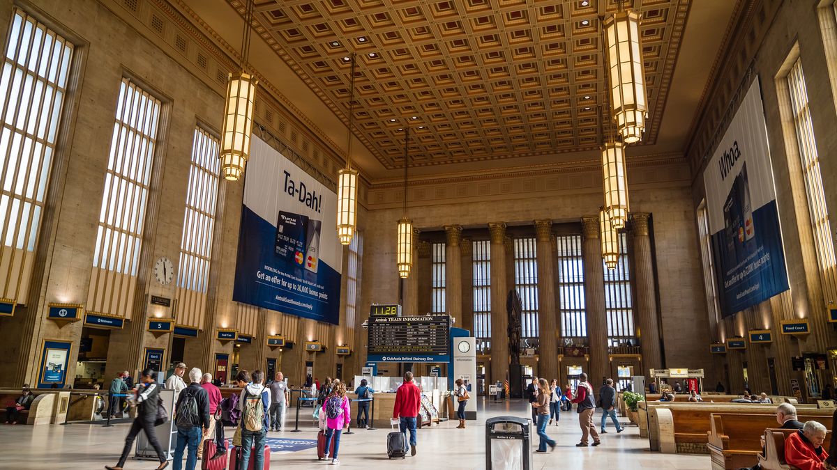 The interior of 30th Street Station in Philadelphia. There are many people. There are light fixtures hanging from the ceiling. The ceiling is high. 