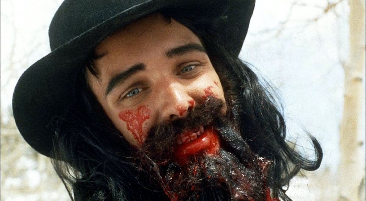 Trey Parker in close-up as Alferd Packer in Cannibal! The Musical, wearing a black Stetson hat, a long black wig, and a long fake black beard with blood smeared thickly across his cheeks, nose, mustache, beard, lips, and teeth