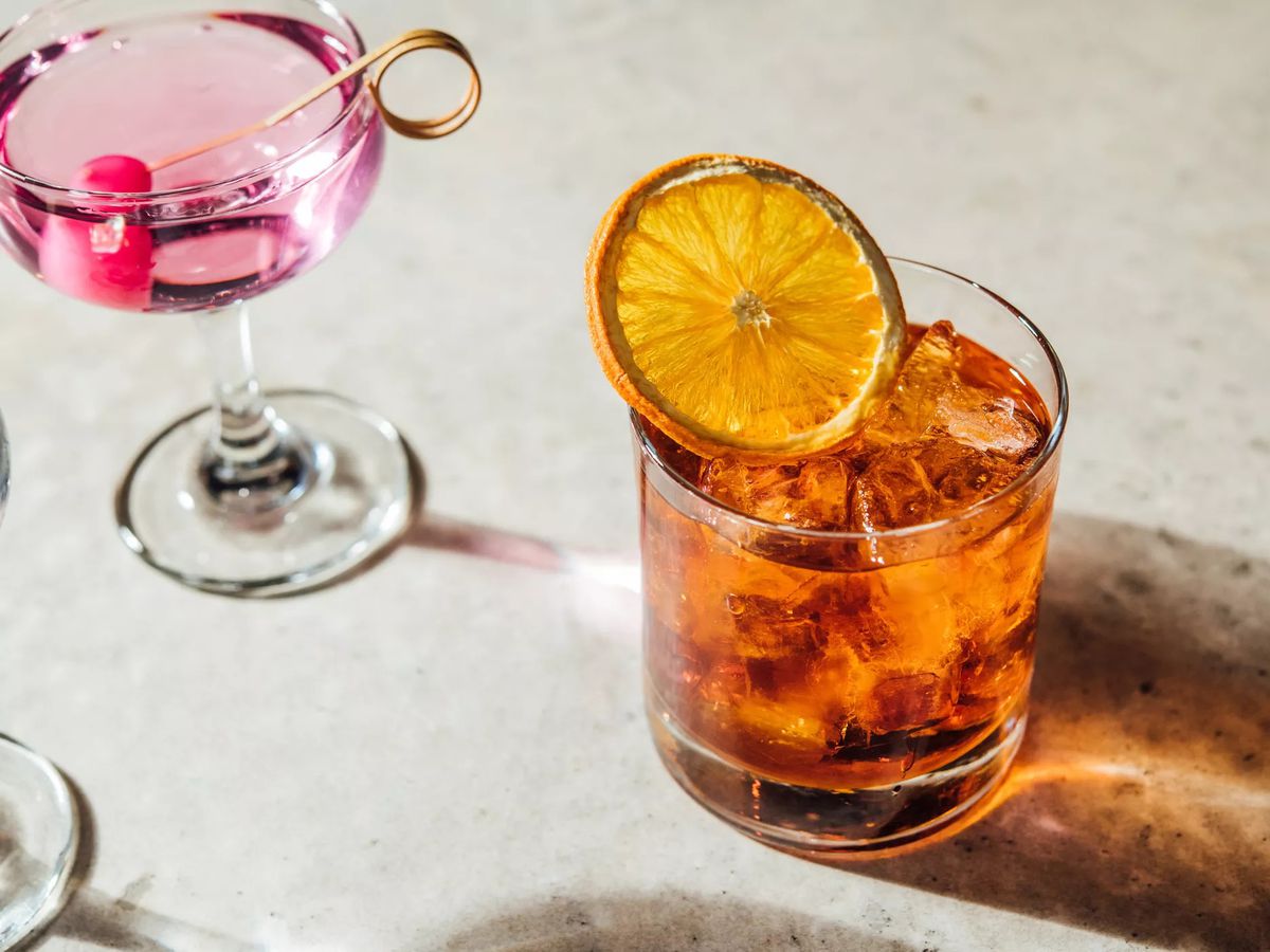 A martini glass is filled with a pink-tinted cocktail and a rocks glass is filled with a dark liquid over ice and topped with a candied orange slice