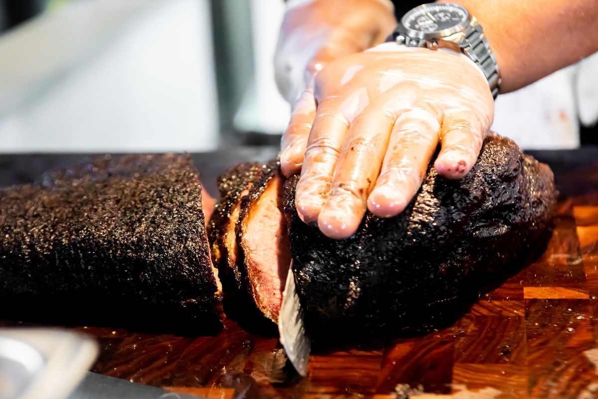 A hand rests on top of a piece of smoked brisket as it’s being sliced