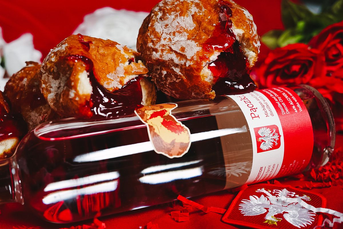Three paczki with raspberry filling behind a clear bottle with red and white packaging and roses in the background.